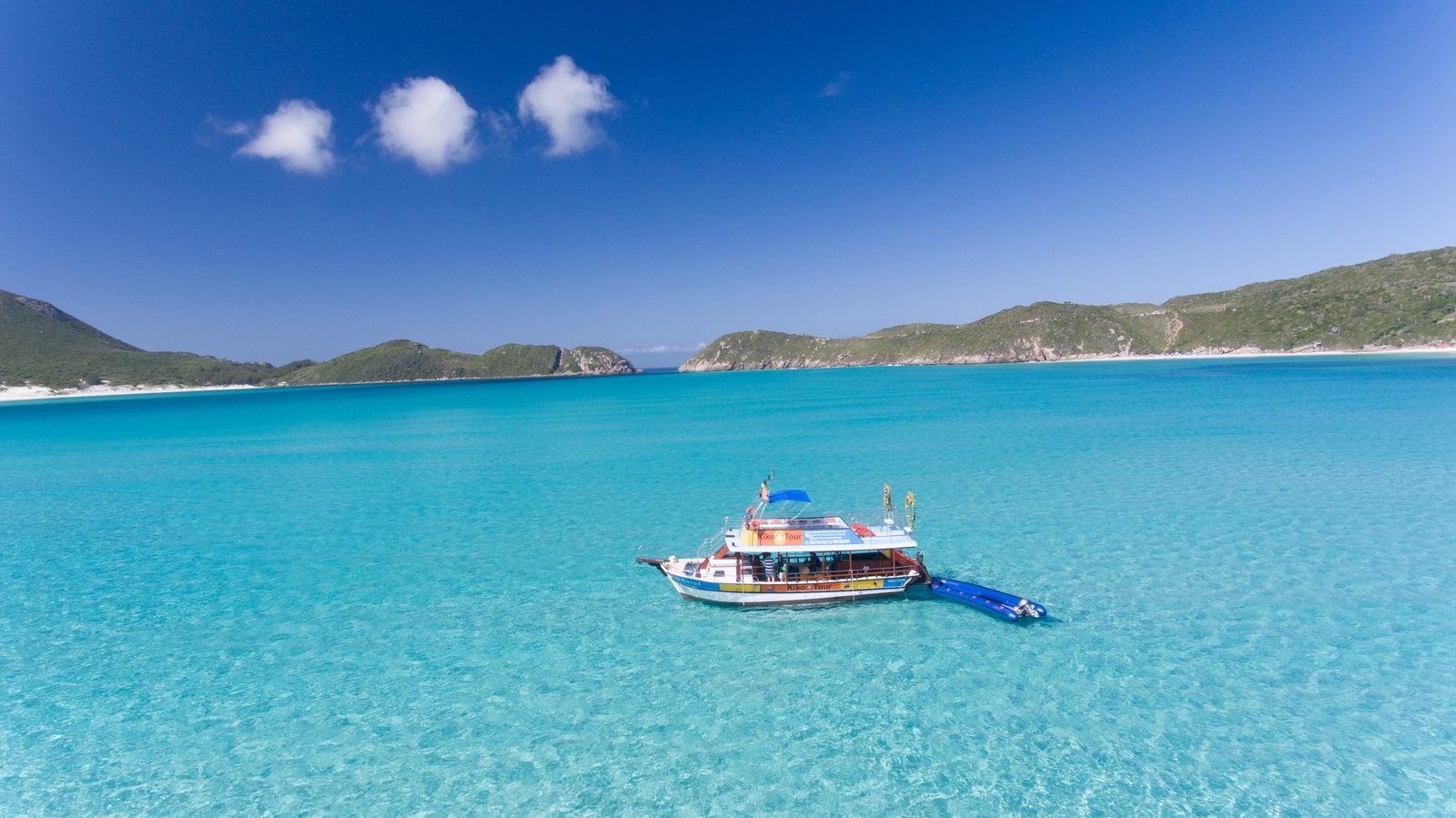 Barco da Kiko Tour ancorado em água turquesa transparente em Arraial do Cabo, com o tobogã inflável ao lado