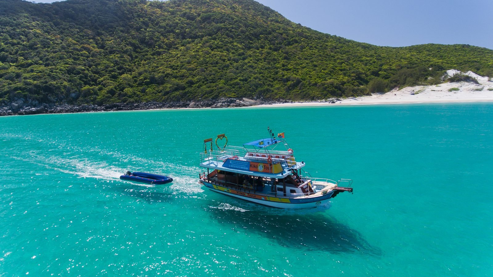 Barco da Kiko Tour com o tobogã inflável diante de uma faixa de areia branca e deserta em Arraial do Cabo